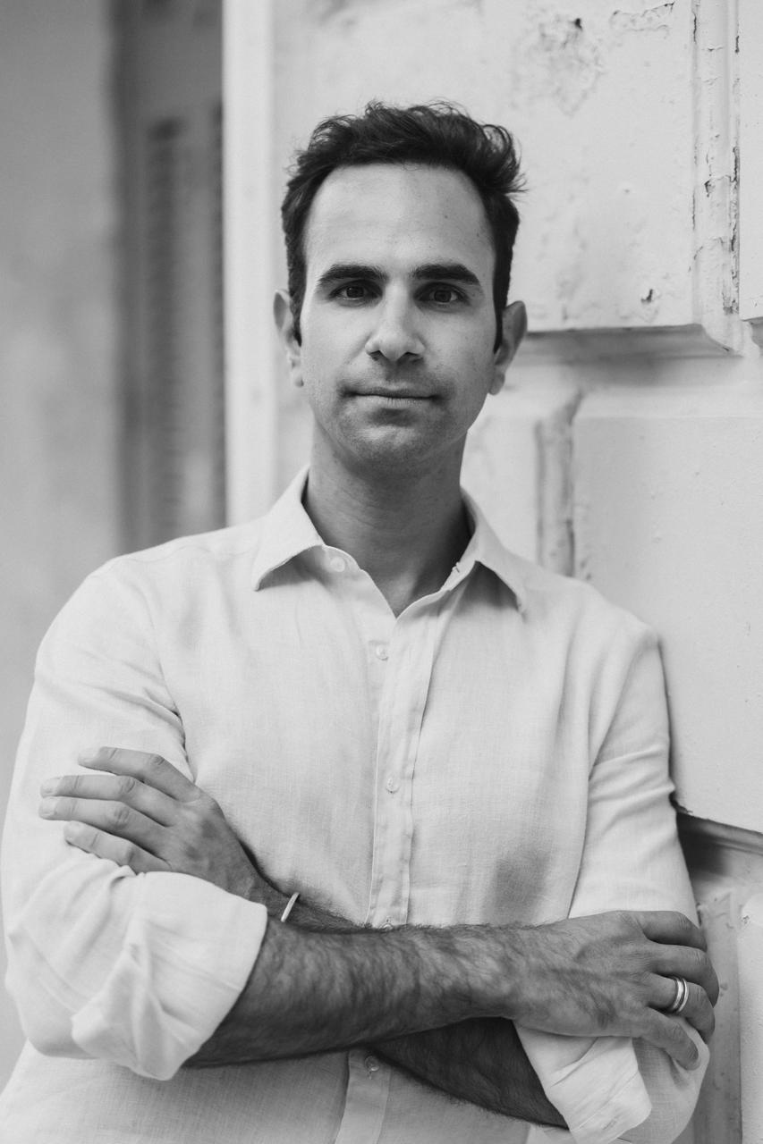 Kyriacos Papademetriou, black and white portrait, arms crossed, leaning on a white wall